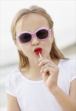 Girl (10-11) wearing sunglasses on beach is licking lollypop. Photo : Momentimages
