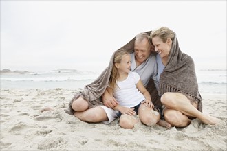 Smiling parents with daughter (10-11) on coastline covered by blanket. Photo : Momentimages