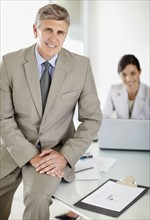 Mature business sitting on desk with secretary in background. Photo : Momentimages