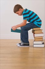 Boy (10-11) sitting on stack of books and reading book. Photo : Daniel Grill