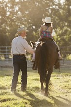 Senior man assisting granddaughter (8-9) horseback riding in ranch. Photo : Mike Kemp