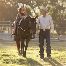 Senior man assisting granddaughter (8-9) horseback riding in ranch. Photo : Mike Kemp