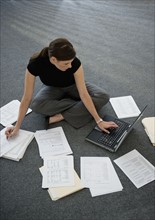Woman sitting on floor with laptop with documents around.