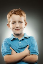 Portrait of smiling redhead boy (4-5) wearing blue polo shirt with arms crossed, studio shot. Photo