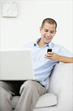 South Africa, Young man sitting on sofa with laptop and texting messages. Photo : momentimages