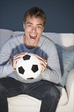 Mid adult man sitting on sofa holding soccer ball and watching tv.