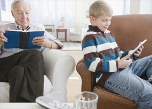 Grandfather reading book, grandson (8-9) reading e-book in living room. Photo : Jamie Grill