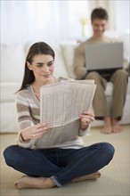Young woman reading newspaper while man is using laptop in background.