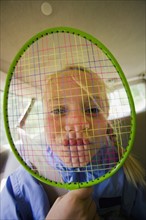 Portrait of girl (6-7) holding badminton racquet over face. Photo : Noah Clayton