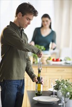 Man opening wine while woman is preparing food in background.