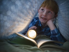 Boy (8-9) reading book under bed covers. Photo : Jamie Grill Photography