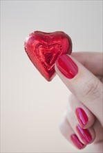 Close up of woman's fingers with red nail polish holding chocolate in shape of heart in red wrapping.