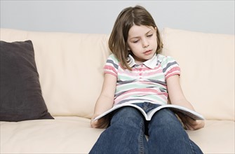 Girl (6-7) reading book on sofa. Photo : Justin Paget