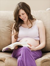 Young pregnant woman sitting on sofa, reading book. Photo : Mike Kemp