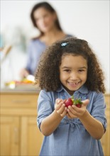 Smiling girl (6-7) holding strawberries with defocused woman in background.