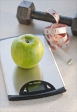 Green apple on weight scale, tape measure and exercise weight in background.