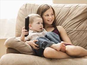 Girl (10-11) with brother (2-3) sitting on sofa, watching TV. Photo : Mike Kemp