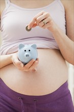 Young pregnant woman putting coin into blue piggybank. Photo: Mike Kemp