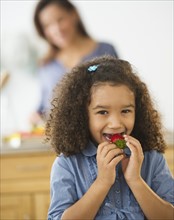 Smiling girl (6-7) eating strawberries with defocused woman in background.