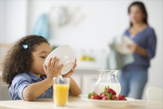 Girl (6-7) eating breakfast with defocused mother in background.