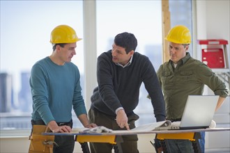 Three man reading blueprint and using laptop on construction site.