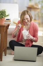 Woman sitting on floor, using laptop and eating take out food.