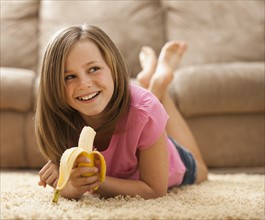 Girl (10-11) lying on rug, eating banana. Photo : Mike Kemp