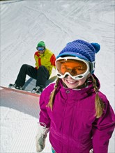 Daughter (10-11) posing with father sitting on ground with snowboard in background . Photo :