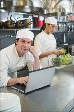 Chef using laptop and cook preparing food in commercial kitchen.