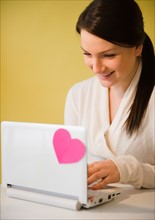 Smiling young woman using laptop with pink heart. Photo : Jamie Grill Photography