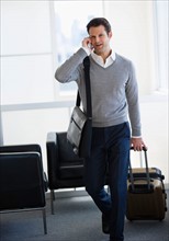 Businessman pulling suitcase and talking on mobile phone in airport lounge.
