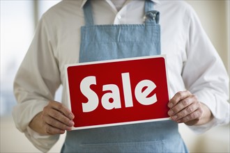 Man wearing apron holding "sale" sign, mid section.