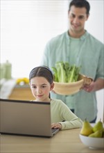 Girl (8-9) using laptop and father preparing food in domestic kitchen.