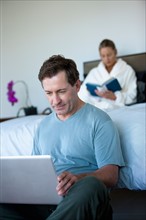 Mature man with laptop in bedroom and woman reading book in bed. Photo : Rob Lewine