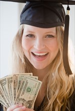 Young woman wearing graduation gown holding banknotes. Photo : Jamie Grill
