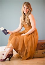 Woman in dress sitting on luggage and holding passport. Photo: Jamie Grill