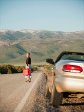 USA, Utah, Kanosh, Woman carrying canister walking along empty road, her car parked on roadside.
