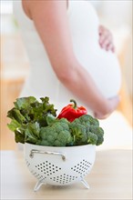 Fresh vegetables in colander, pregnant woman in background.