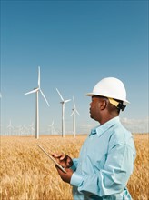 USA, Oregon, Wasco, Engineer standing in wheat field in front of wind turbines, using digital