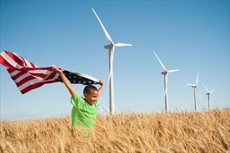 USA, Oregon, Wasco, Boy (8-9) flying american flag in wheat field with wind turbines in background.