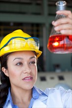 Woman wearing lab coat and hardhat looking at red liquid in beaker. Photo: db2stock