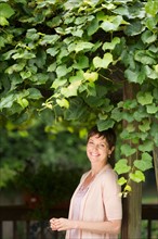 Portrait of mature woman leaning against wooden post overgrown with ivy.