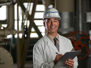 Man in lab coat and hard hat holding clipboard. Photo : db2stock