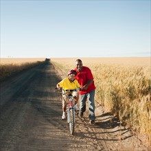 Father teaching son (8-9) how to cycle on along dirt road.