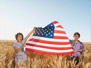 Girls (10-11, 12-13) holding american flag in wheat field.