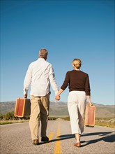 Mid adult couple walking along empty road carrying suitcases.