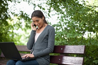 Young woman sitting on park bench using laptop and cell phone. Photo: Jan Scherders