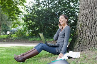 Young woman sitting under tree using laptop and cell phone. Photo: Jan Scherders