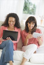 Two women sitting on sofa using digital tablets.