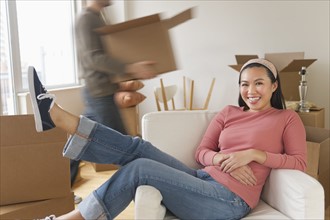 Man carrying boxes, woman resting in chair at new home.